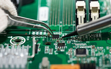 Electronic component  held with tweezers by an engineer over a green motherboard.